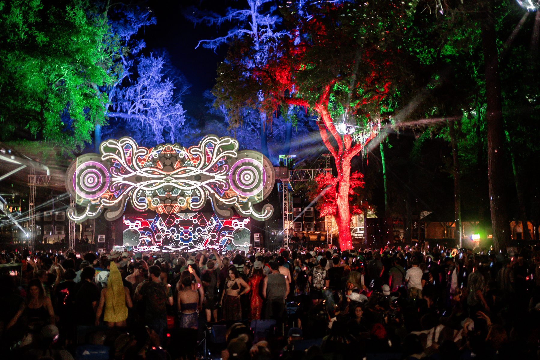 Festival stage in the woods with a lit art structure and crowd at night